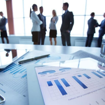 Fotografia com foco em planilhas e um tablet com gráficos financeiros sobre a mesa, exibindo ao fundo o reflexo e a silhueta de executivos conversando integrados em um escritório, ilustrando a visão global da comunicação sistêmica nas empresas.