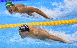 Fotografia de dois nadadores profissionais competindo em uma piscina durante a prova de nado borboleta, com o atleta em primeiro plano demonstrando foco extremo, ilustrando o conceito de desenvolver confiança, disciplina e alta performance.