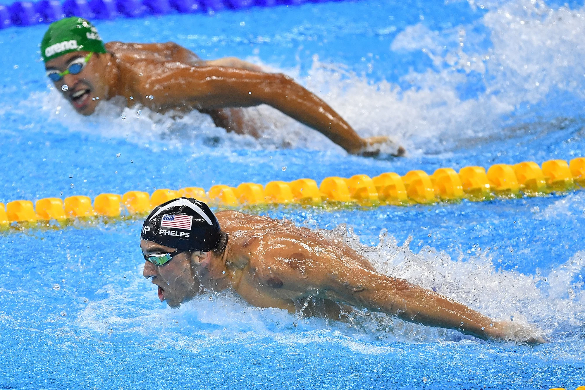 Fotografia de dois nadadores profissionais competindo em uma piscina durante a prova de nado borboleta, com o atleta em primeiro plano demonstrando foco extremo, ilustrando o conceito de desenvolver confiança, disciplina e alta performance.